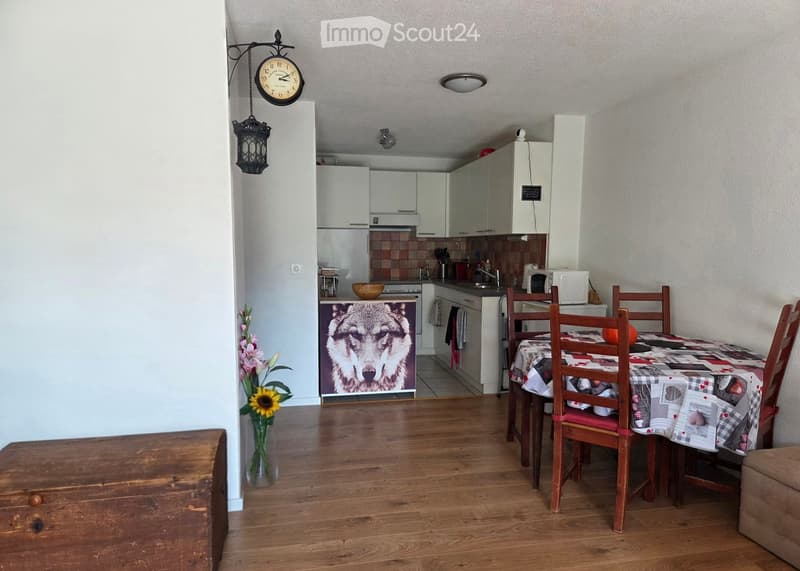 A well-lit dining and kitchen area with wooden flooring, a dining table with chairs, a vase with flowers, a clock, and a kitchen with cabinets, a refrigerator, microwave, and various appliances.