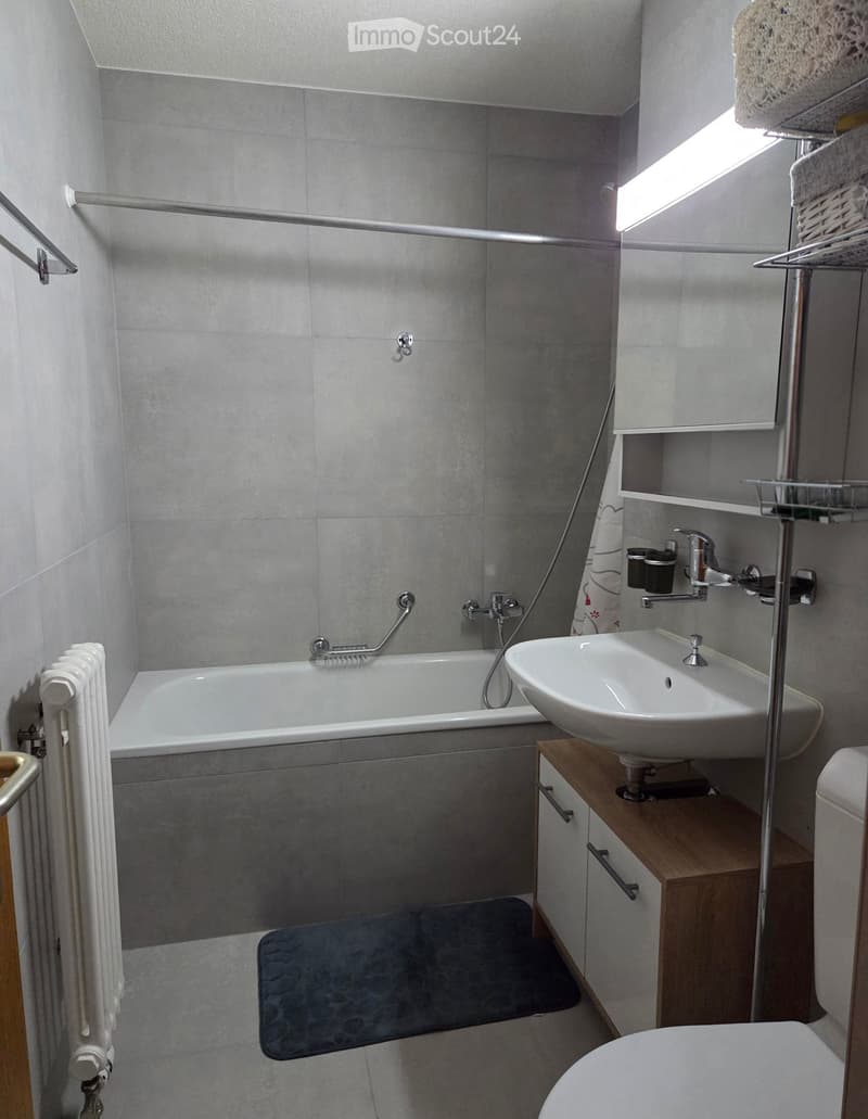Modern bathroom with a bathtub, sink, toilet, radiator, and a mirror. The walls are tiled in grey, and there is a wooden shelf beneath the sink.