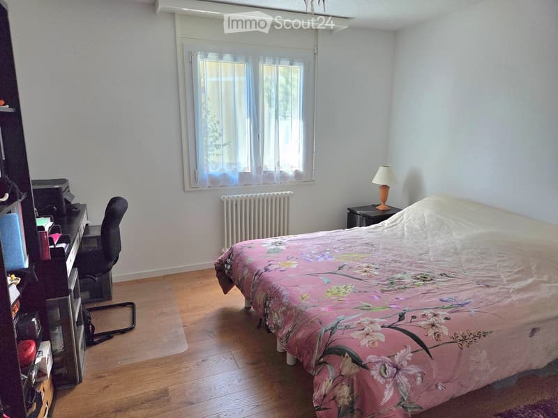 A well-lit bedroom with wooden flooring, a large window with sheer curtains, a bed with a pink floral duvet, a desk with a chair, a small table with a lamp, and a radiator below the window.