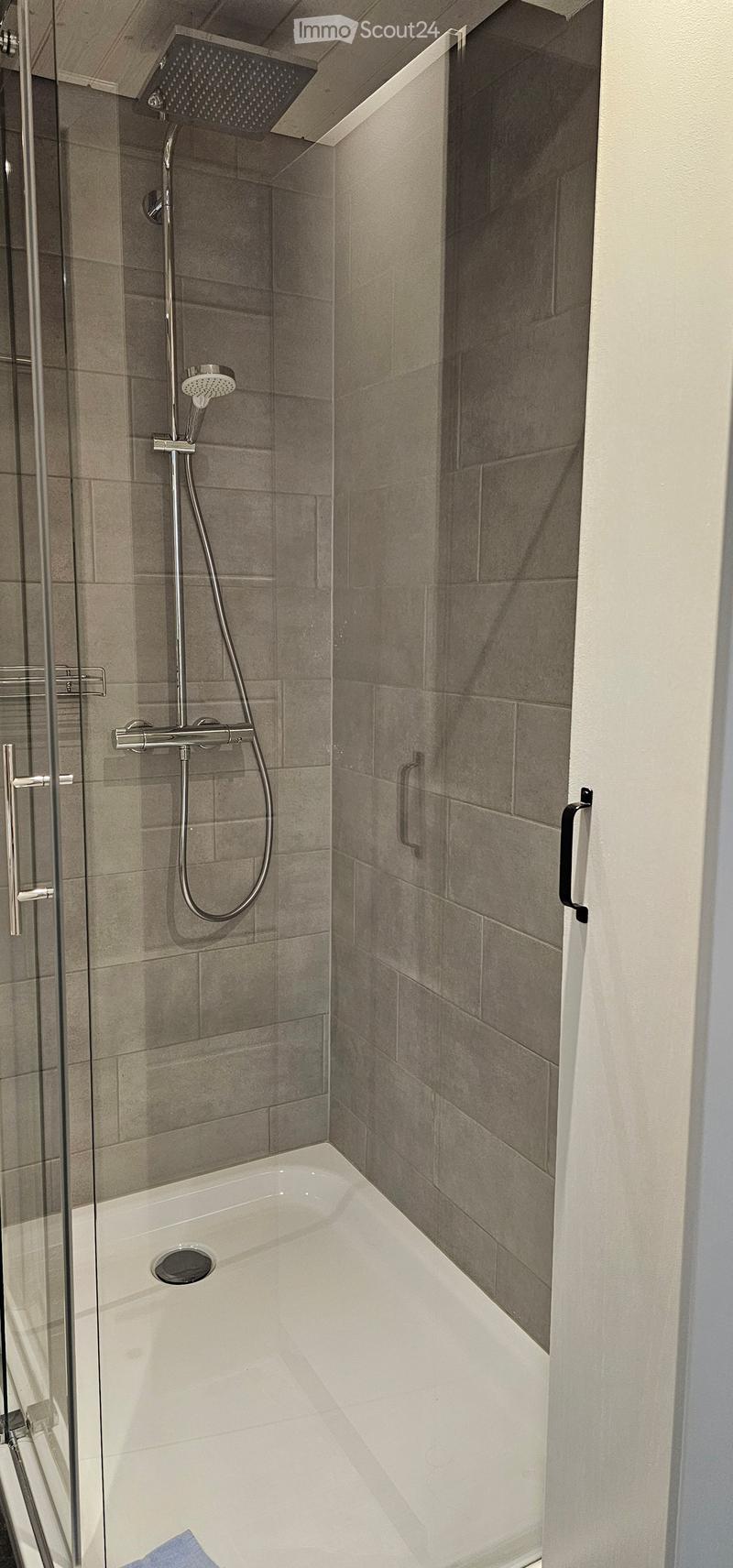 Modern bathroom with a bathtub, glass shower enclosure, and a handheld showerhead. The walls are tiled in a neutral gray color, and the shower features a sleek, contemporary design.