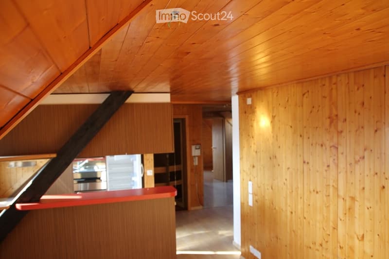 A modern wooden hallway with a slanted wooden ceiling, warm lighting, and a red counter in the foreground. The hallway leads to another room with wooden walls and a visible staircase.