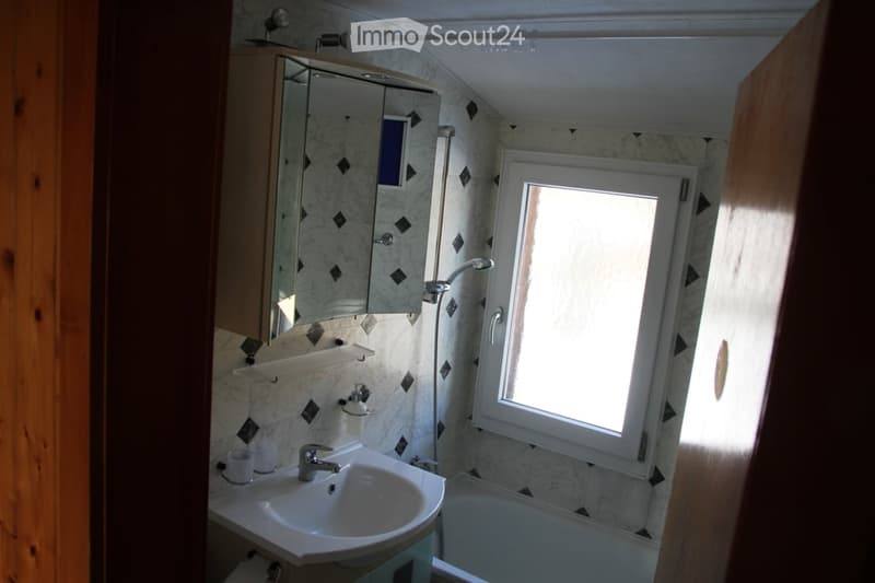 Bathroom with a sink, mirror, bathtub, and showerhead. The walls are tiled with a diamond pattern, and there is a window letting in natural light.