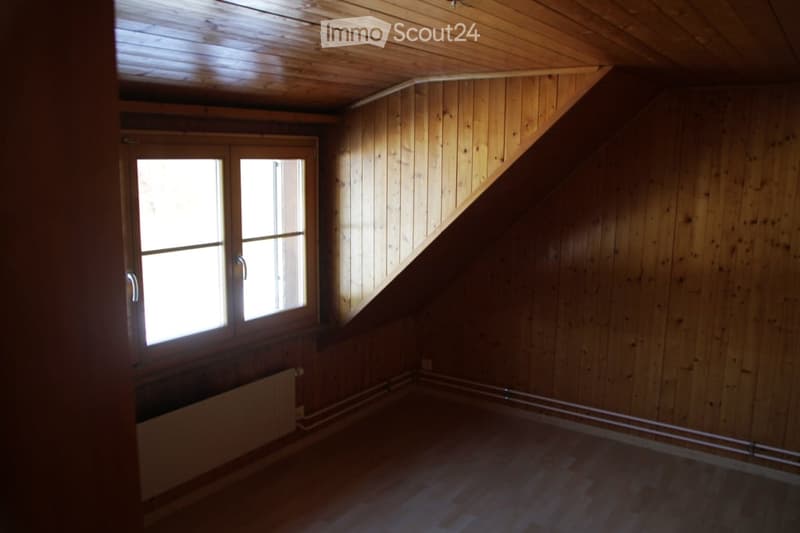 Empty attic room with wooden walls, ceiling, and floor, featuring a large window and radiator.