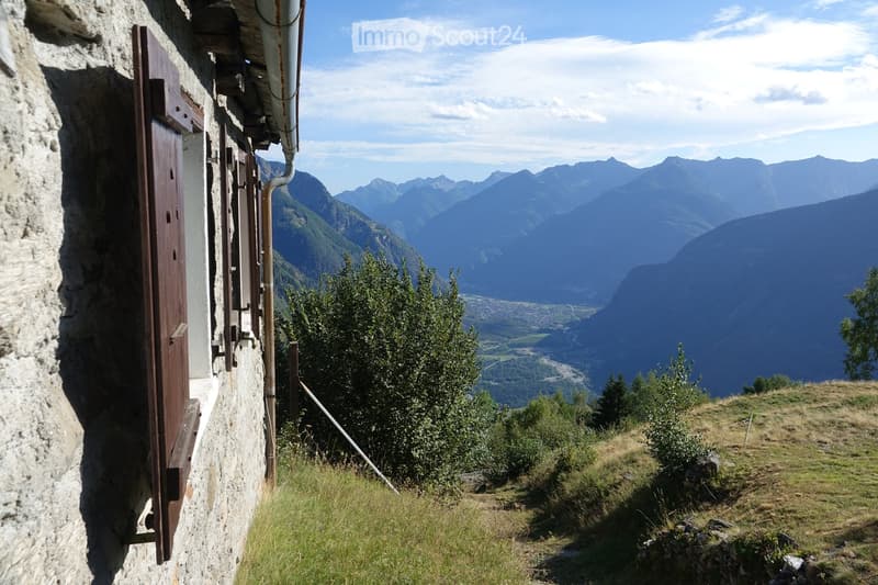A rustic stone building with a wooden door and metal pipes, situated on a grassy slope with a panoramic mountain view in the background.
