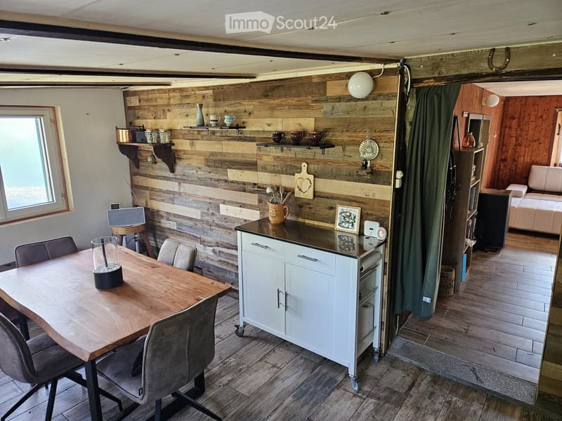 A cozy, rustic kitchen and living area with wooden walls, a dining table with chairs, a white kitchen cabinet, and a small living space with a sofa.