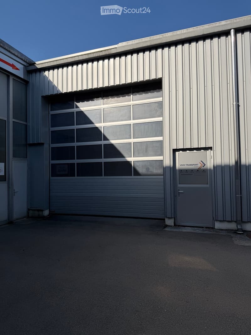 A modern industrial building with a large garage door and a smaller side entrance. The structure features a flat, metallic roof and large windows above the garage door.