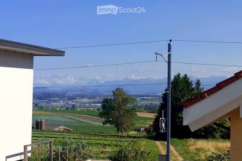View from a house balcony showing a rural landscape with fields, trees, and distant mountains.