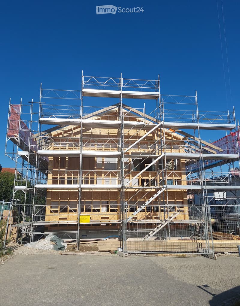 A two-story house under construction, with scaffolding and exposed wooden framing, surrounded by a metal fence and construction materials.