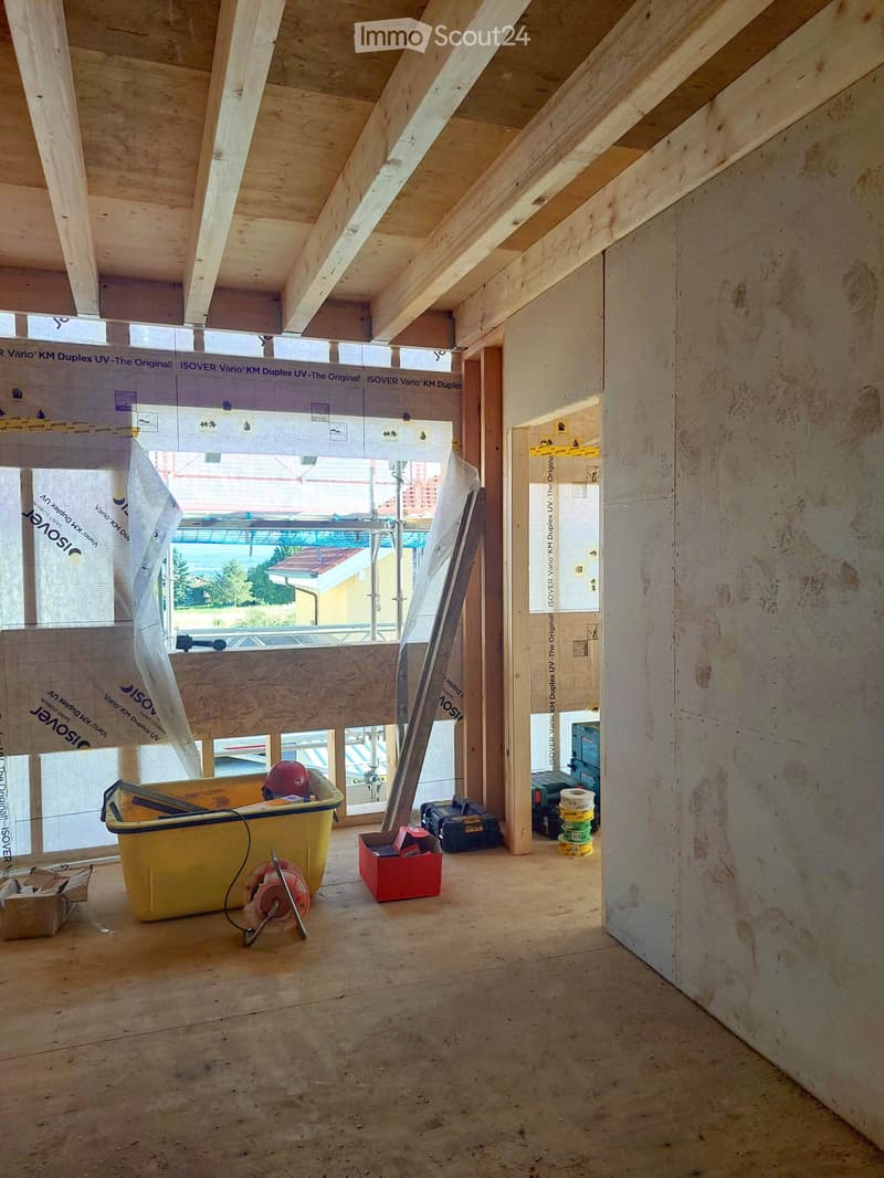 Empty room with exposed wooden beams and unfinished walls, featuring a large window with protective covering, construction materials, and tools scattered around.