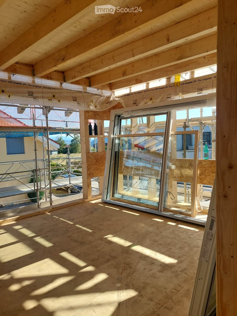 Empty room with exposed wooden beams and a large window, showing natural light and shadows on the floor.