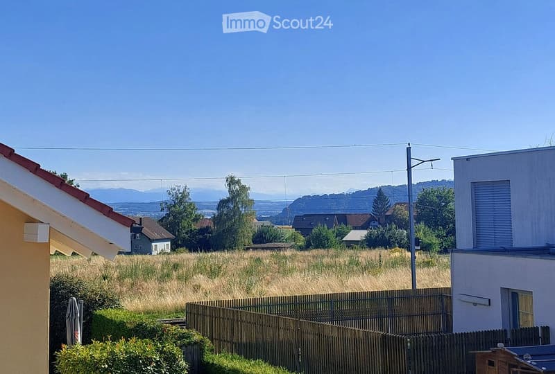 View of a rural landscape with a wooden fence, tall grass field, and distant mountains under a clear blue sky.
