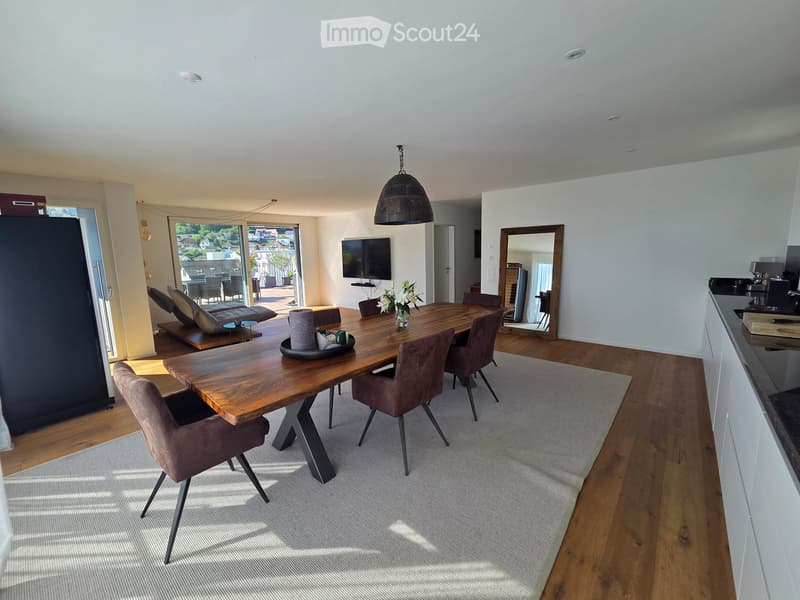 Modern dining area with a wooden dining table, brown chairs, a hanging pendant light, a television mounted on the wall, and a view of an outdoor patio.