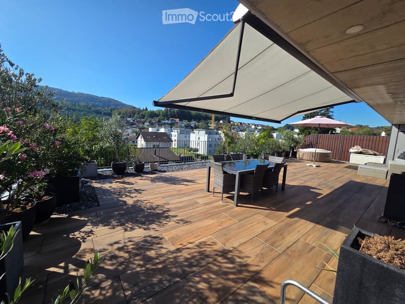 Wooden deck with a dining table, chairs, umbrella, hot tub, and potted plants, overlooking a scenic town and mountains.