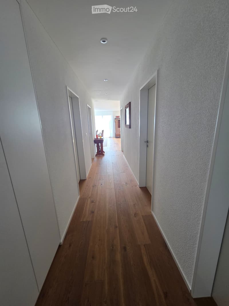 A long, narrow hallway with wooden flooring and white walls, featuring several doors and a small table with items at the end.