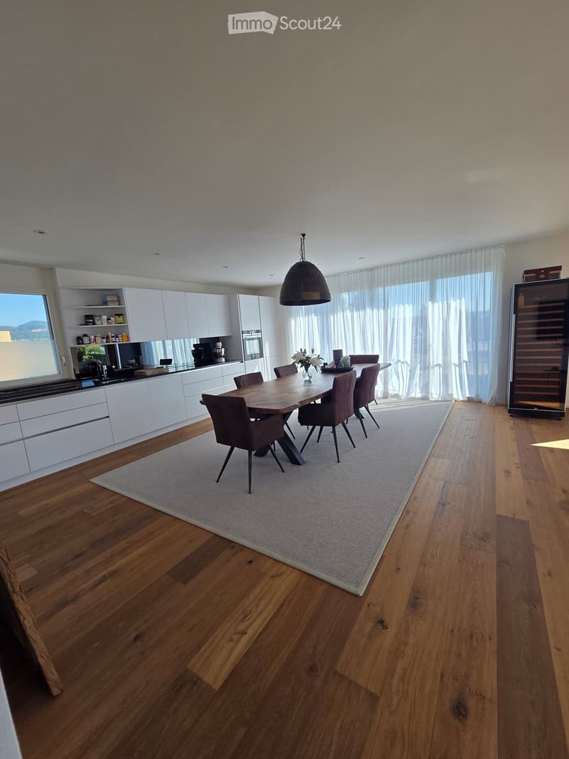Open-plan kitchen and dining area with white cabinets, wooden dining table, six chairs, pendant light, and large windows with curtains.