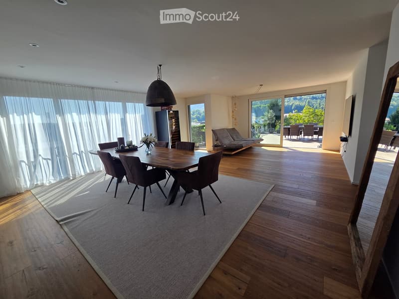 Bright dining area with wooden floor, large windows, dining table with chairs, sofa, and pendant light. View of outdoor patio with seating.