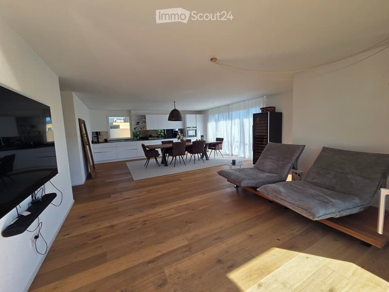 Open-plan living and dining area with wooden flooring, a grey sofa, a dining table with chairs, a modern kitchen, and a wine rack.