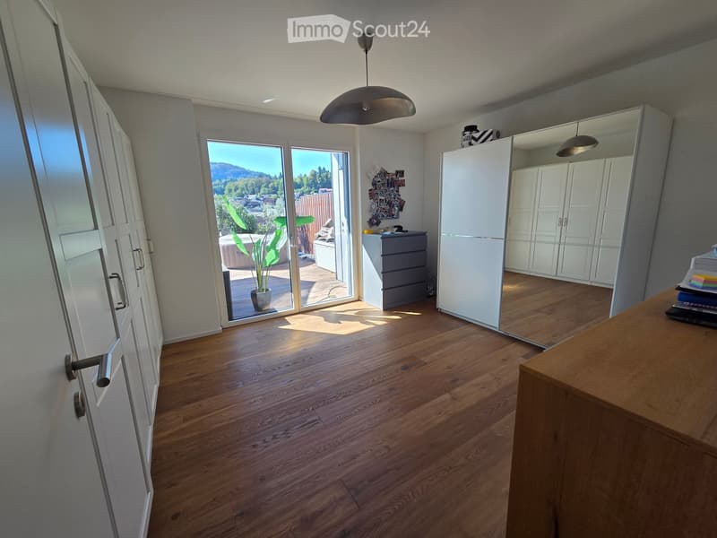 Bright, modern room with wooden flooring, large sliding glass doors leading to a balcony, wardrobe, small table with laptop, and a potted plant visible through the glass doors.