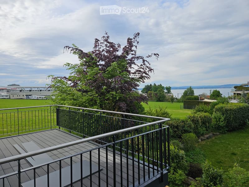 Balcony with wooden decking and metal railing, overlooking a green lawn, trees, and a body of water in the distance.
