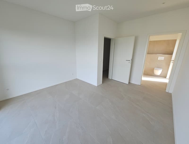 Empty room with light grey tiled flooring and white walls, featuring two open doors leading to other spaces, including a bathroom with a toilet visible.