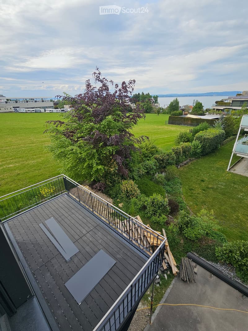 Roof terrace with wooden decking, metal railing, and surrounding greenery, overlooking a grassy area and distant water.