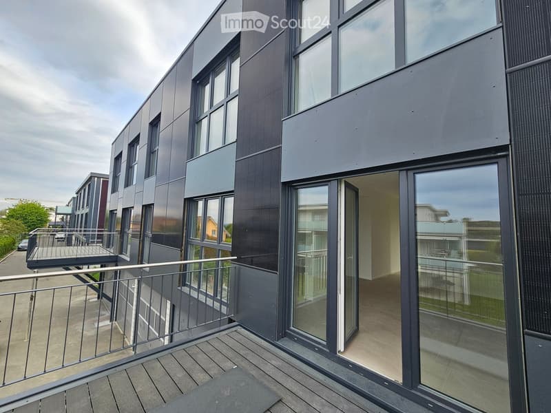 Modern balcony with wooden decking, metal railing, and large glass sliding doors leading to a contemporary building with grey and black facade.