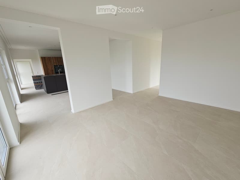 Empty hallway with light-colored tiled flooring and white walls, leading to a kitchen area with dark cabinets and a microwave.