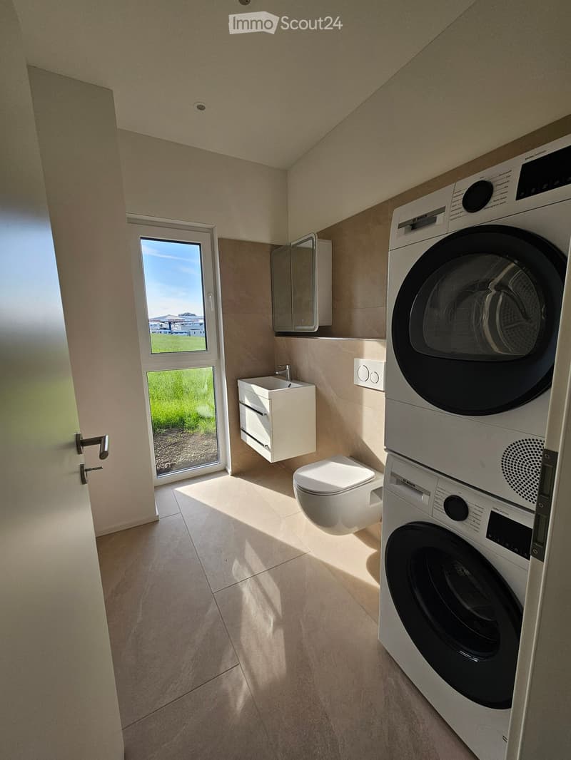 Modern bathroom with a toilet, sink, mirror, and a stacked washing machine/dryer unit. Natural light streams in from a large window.
