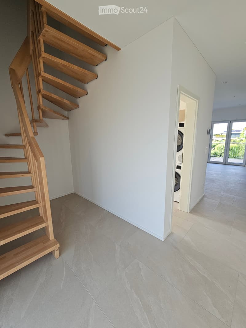 Modern hallway with light-colored tiled flooring, white walls, and a wooden staircase leading upwards. A doorway leads to another room with a washing machine visible.