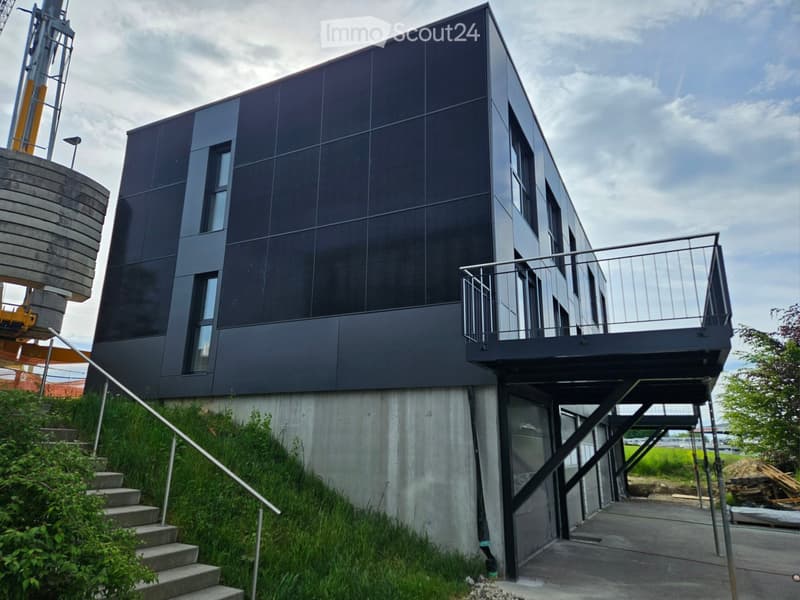 Modern two-story building with dark grey facade, large windows, and a raised ground floor accessed by concrete steps.