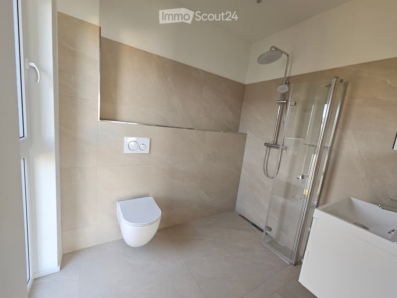 Modern bathroom with beige tiled walls and floor, featuring a toilet, a glass-enclosed shower with a handheld showerhead, and a white sink with a cabinet.