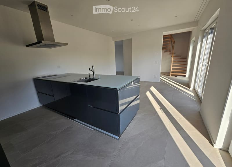Modern kitchen with a dark grey island, stainless steel sink, and a range hood. The space features large windows and a visible staircase leading to another level.