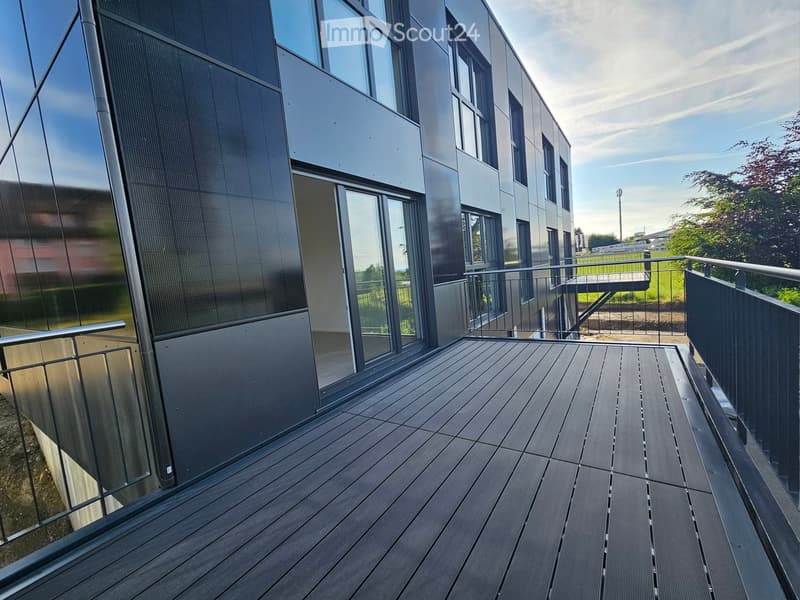 Modern balcony with wooden decking, metal railings, and large windows overlooking a green area.