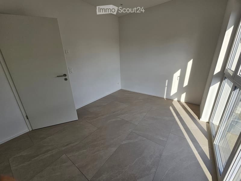 Empty room with tiled floor, white walls, and large windows allowing natural light to enter. The room features a door on one side and is completely unfurnished.