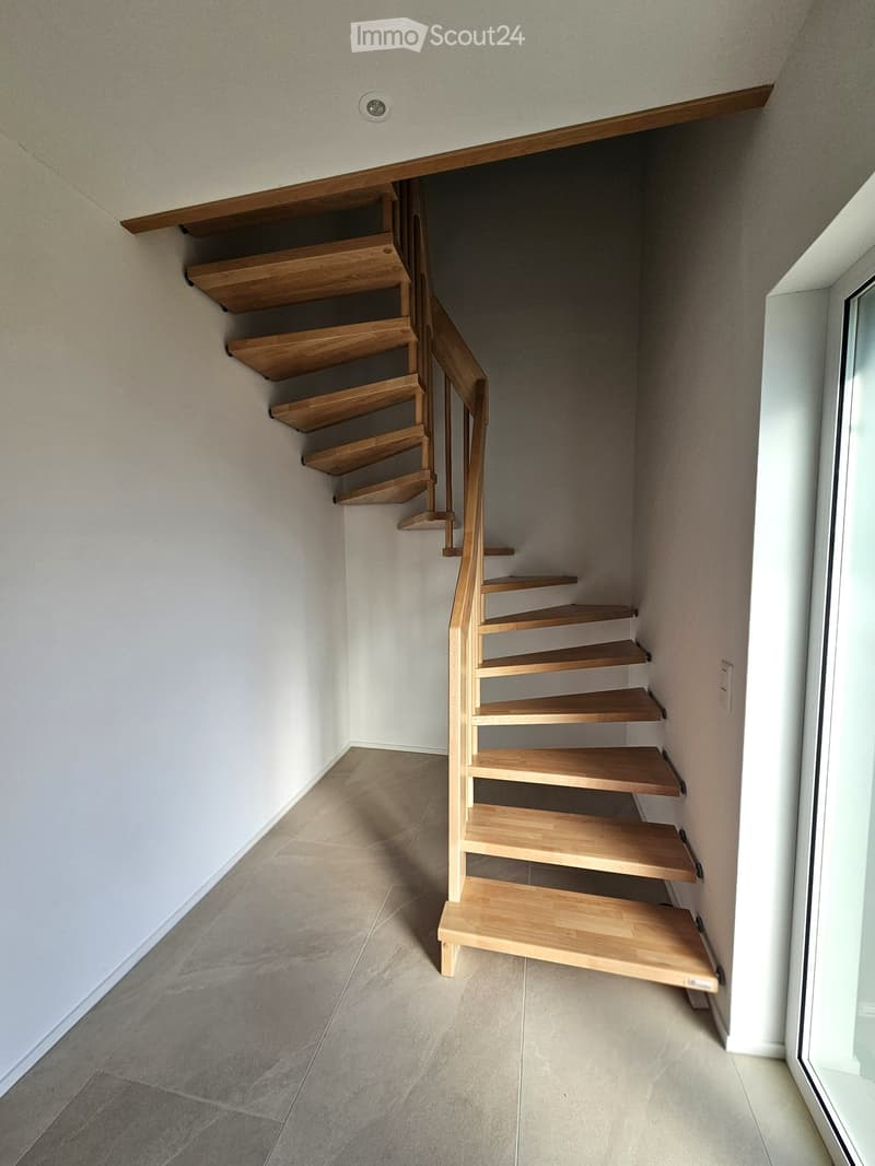 A modern, minimalist staircase with wooden steps and a sleek handrail, set against light-colored walls and a tiled floor.