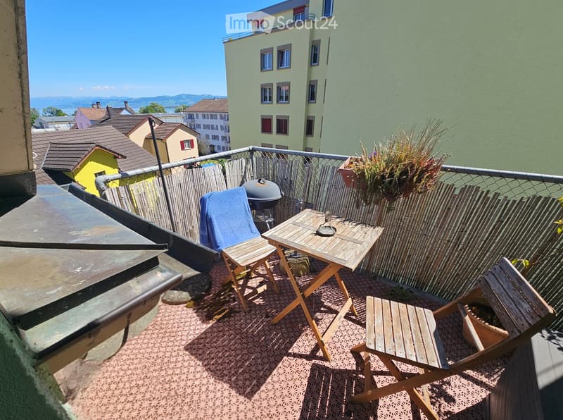 Balcony with wooden table, chairs, grill, and potted plants, overlooking a cityscape with buildings and mountains.