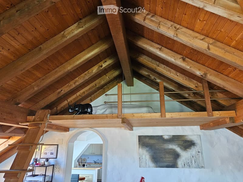 Loft-style room with exposed wooden beams and a raised platform, featuring a shelf with a statue, a mirror, a framed picture, a small table with a chair, and a visible kitchen area with a counter, sink, and faucet.