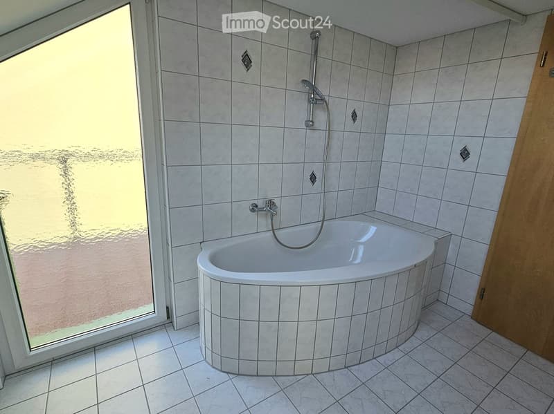 Modern bathroom with a corner bathtub, tiled walls and floor, and a large window offering a view of the outdoor area.