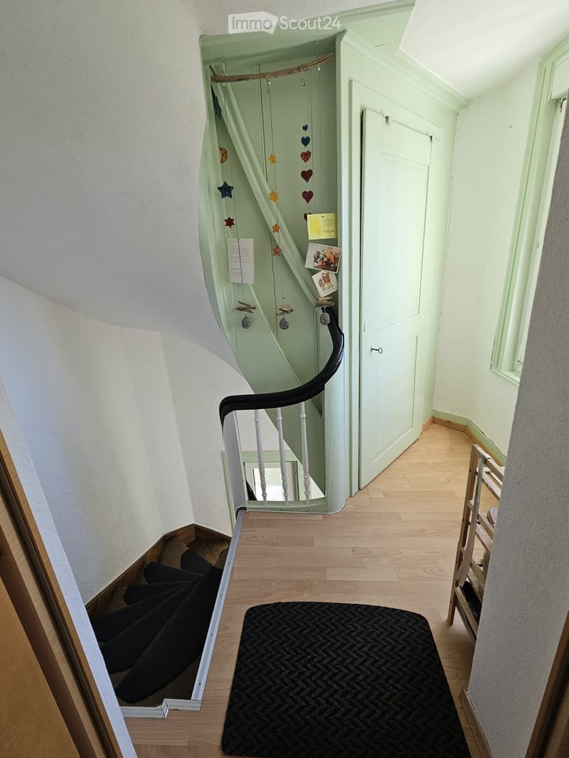 A narrow hallway with wooden flooring, a black runner rug, a staircase with a white railing, and a door on the right side.