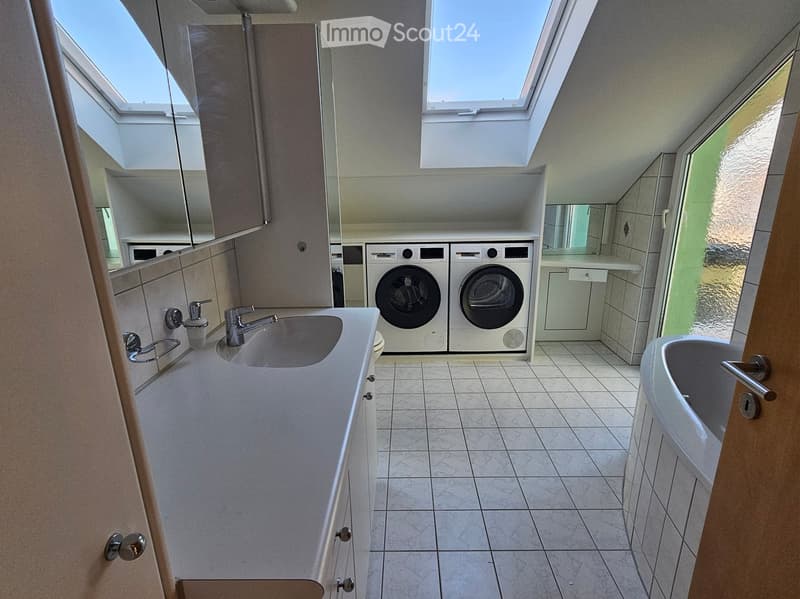 Modern bathroom with a sink, bathtub, washing machine, and dryer. Features include a large mirror, tiled floor, and skylight windows.