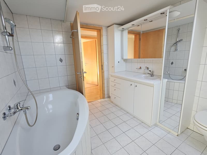Modern bathroom with a bathtub, sink, mirror, and toilet. The walls are tiled, and there is a wooden door leading to another room.