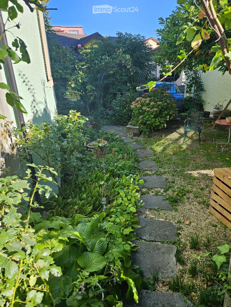A small, well-maintained garden with a stone pathway, various plants, and a small seating area with a table and chair.