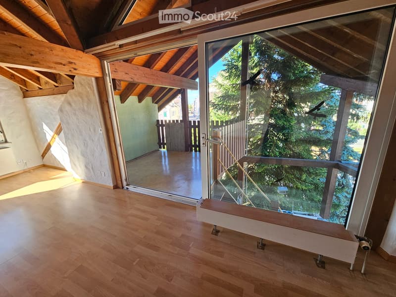 Bright, empty room with wooden flooring and large glass doors leading to a balcony or terrace. The room features exposed wooden beams on the ceiling and a modern, minimalist design.