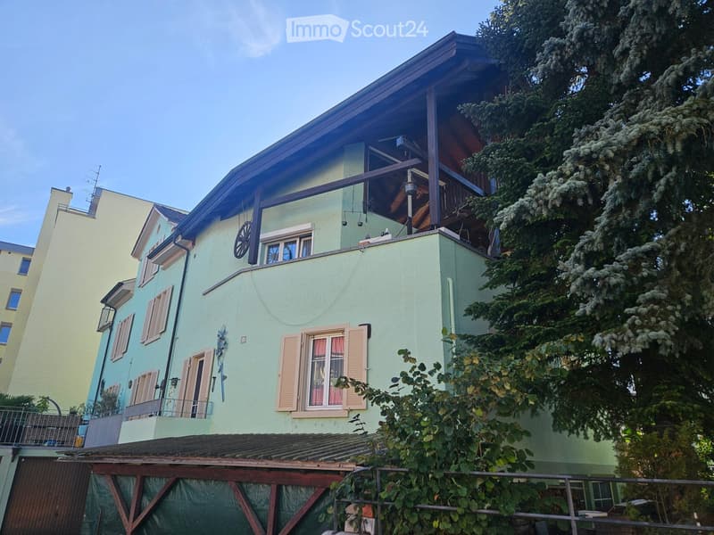 A two-story house with light green walls, multiple windows with shutters, and a wooden balcony on the upper level. The roof is dark, and there is a covered patio area with wooden beams and a small garden in front.