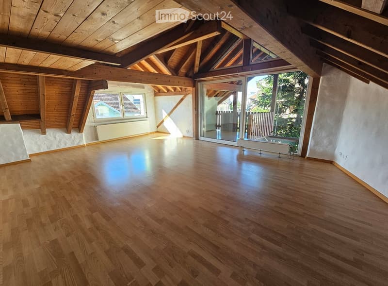 Empty attic room with wooden floors, exposed wooden beams, large windows, and a sliding glass door leading to a balcony.