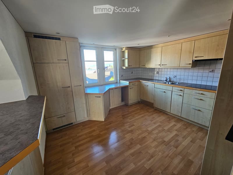 Modern kitchen with light wood cabinets, a central island, and a sink area. The floor is made of polished wood, and the space is well-lit with natural light from a large window.