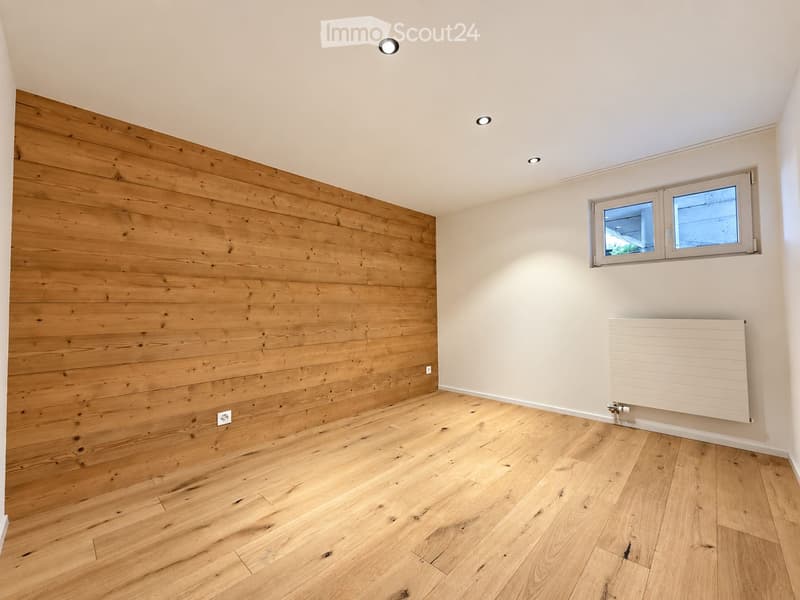 Empty room with wooden flooring and a wooden accent wall, featuring a window and a radiator.