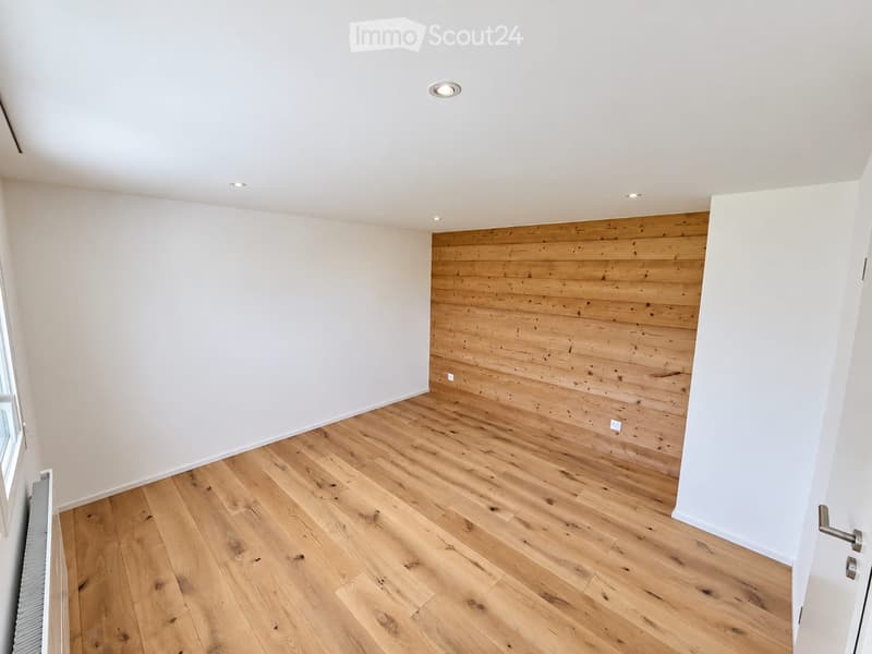 Empty room with light wooden flooring, white walls, and a wooden accent wall. Features recessed ceiling lights and a window on the left side.