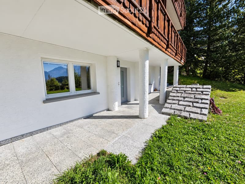 A modern white house with a wooden balcony, large windows, and a stone pathway leading to the entrance, surrounded by greenery and a scenic mountain view.
