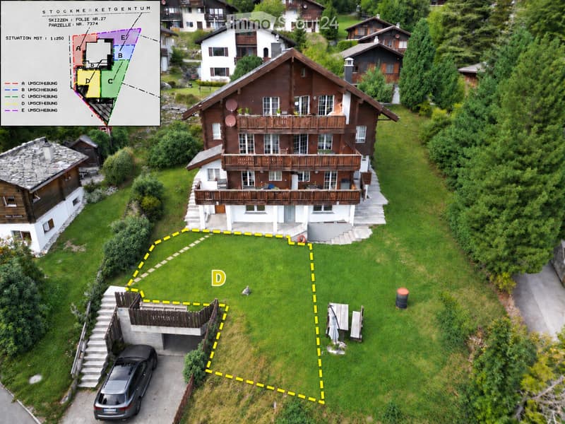 Aerial view of a two-story wooden house with a large balcony, surrounded by a well-maintained lawn and neighboring houses. A car is parked in the driveway, and a small garden area is visible.